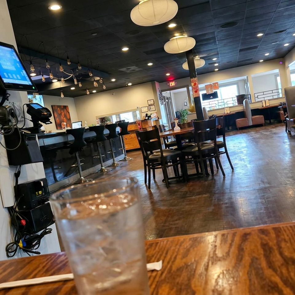 Spacious restaurant dining room with wooden tables, booth seating, pendant lights, a bar counter, and iced water glasses in the foreground.
